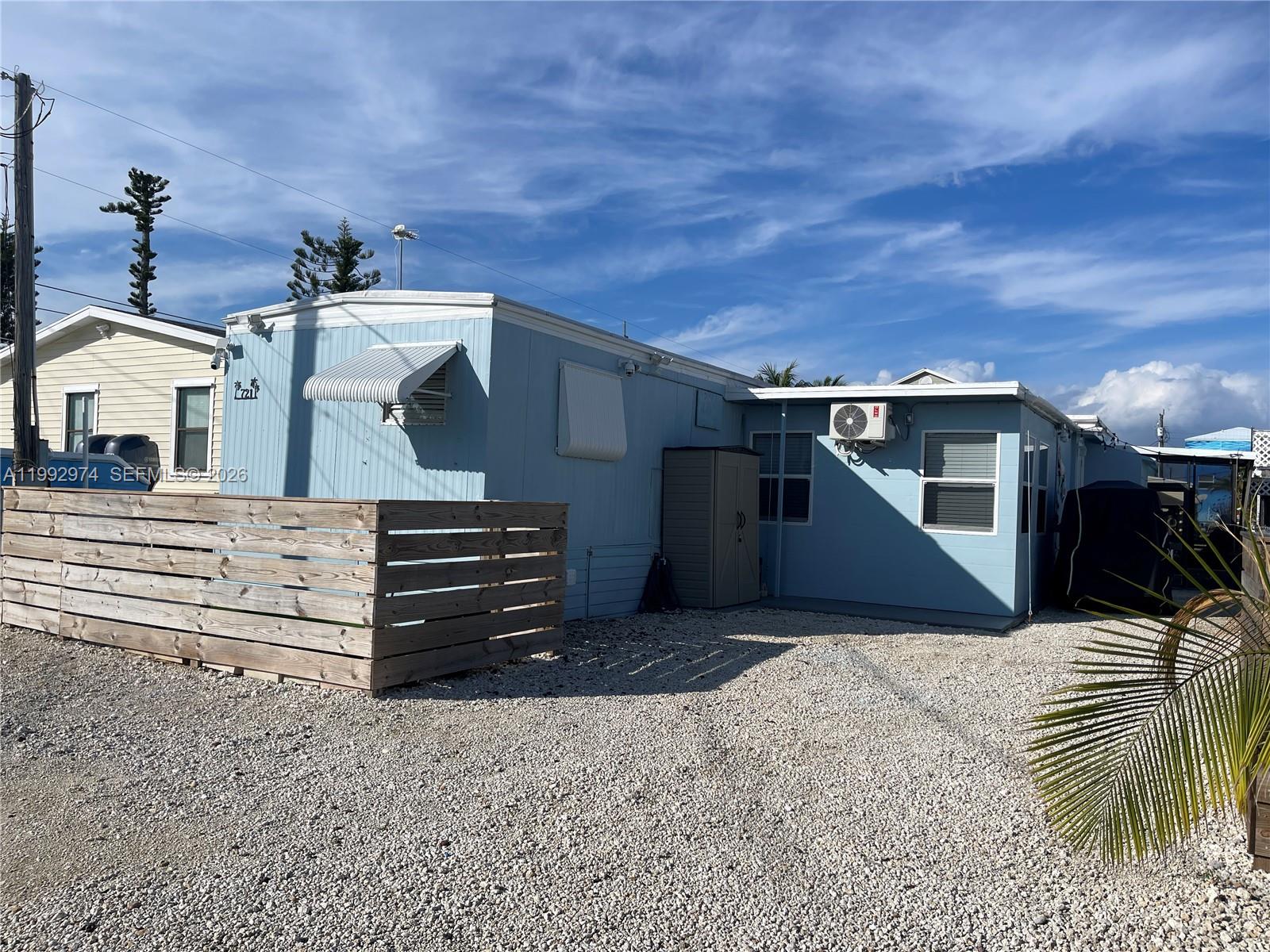 A light blue mobile home features a wooden privacy fence and a gravel driveway — 721 Garden State Ln, Key Largo FL 33037, KEY LARGO, FL
