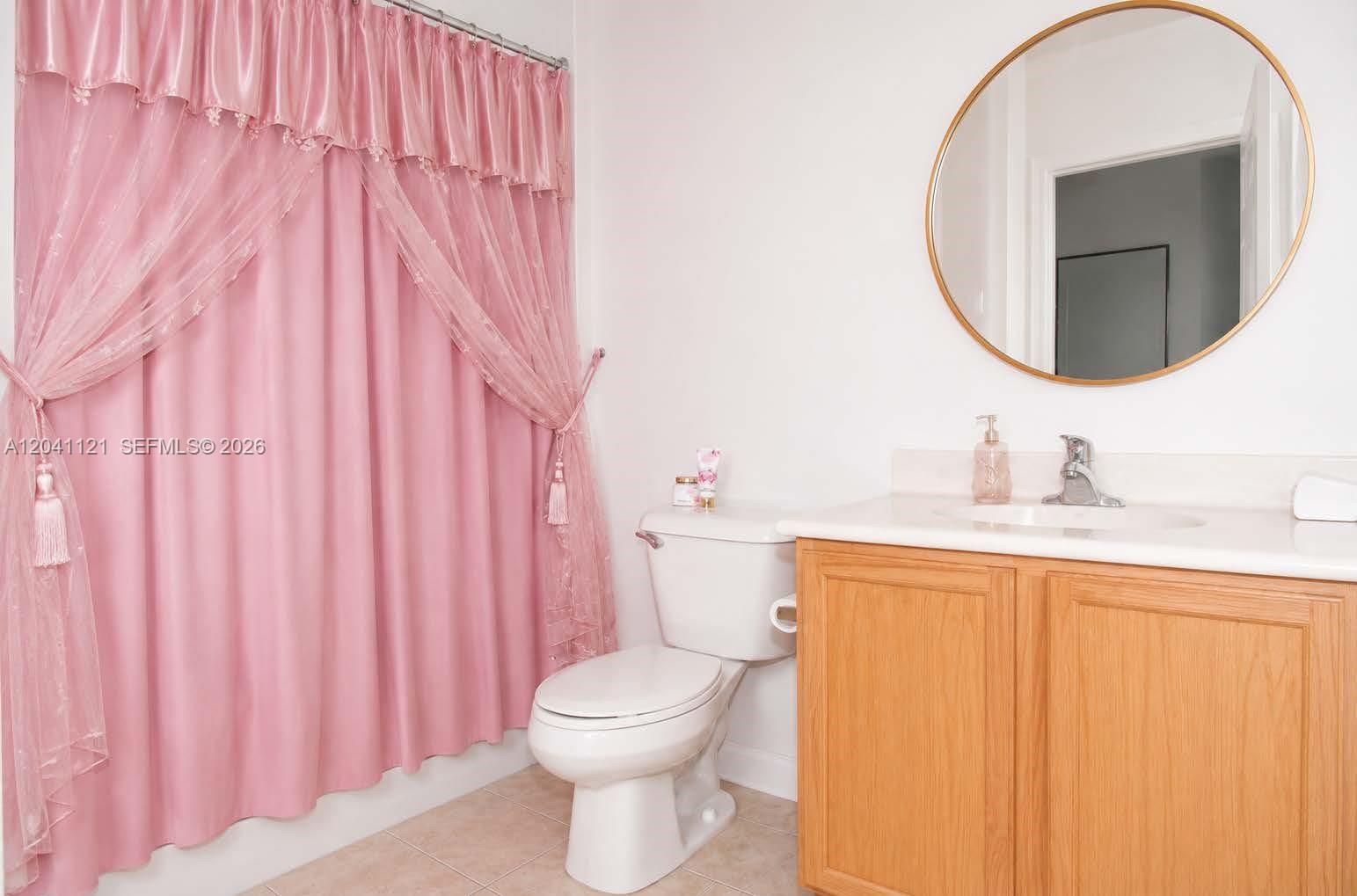 A bathroom features a pink curtain, a white toilet, and a wooden vanity with mirror. k — 13127 SW 44th St # 12202, Miramar FL 33027, MIRAMAR, FL
