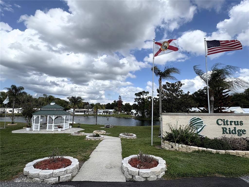 A gazebo sits beside a pond near a Citrus Ridge community sign and two flags — 446 CITRUS RIDGE, DAVENPORT, FL