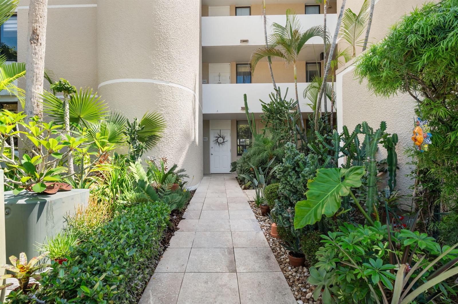 A tiled walkway leads to a white front door between lush tropical landscaping and cacti — 3412 NE Causeway Boulevard # 102, Jensen Beach FL 34957, JENSEN BEACH, FL