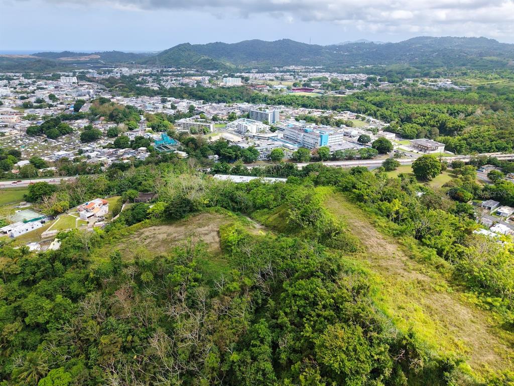 An aerial view shows a grassy hillside in the foreground overlooking a town and mountains — MABO, HUMACAO, PR