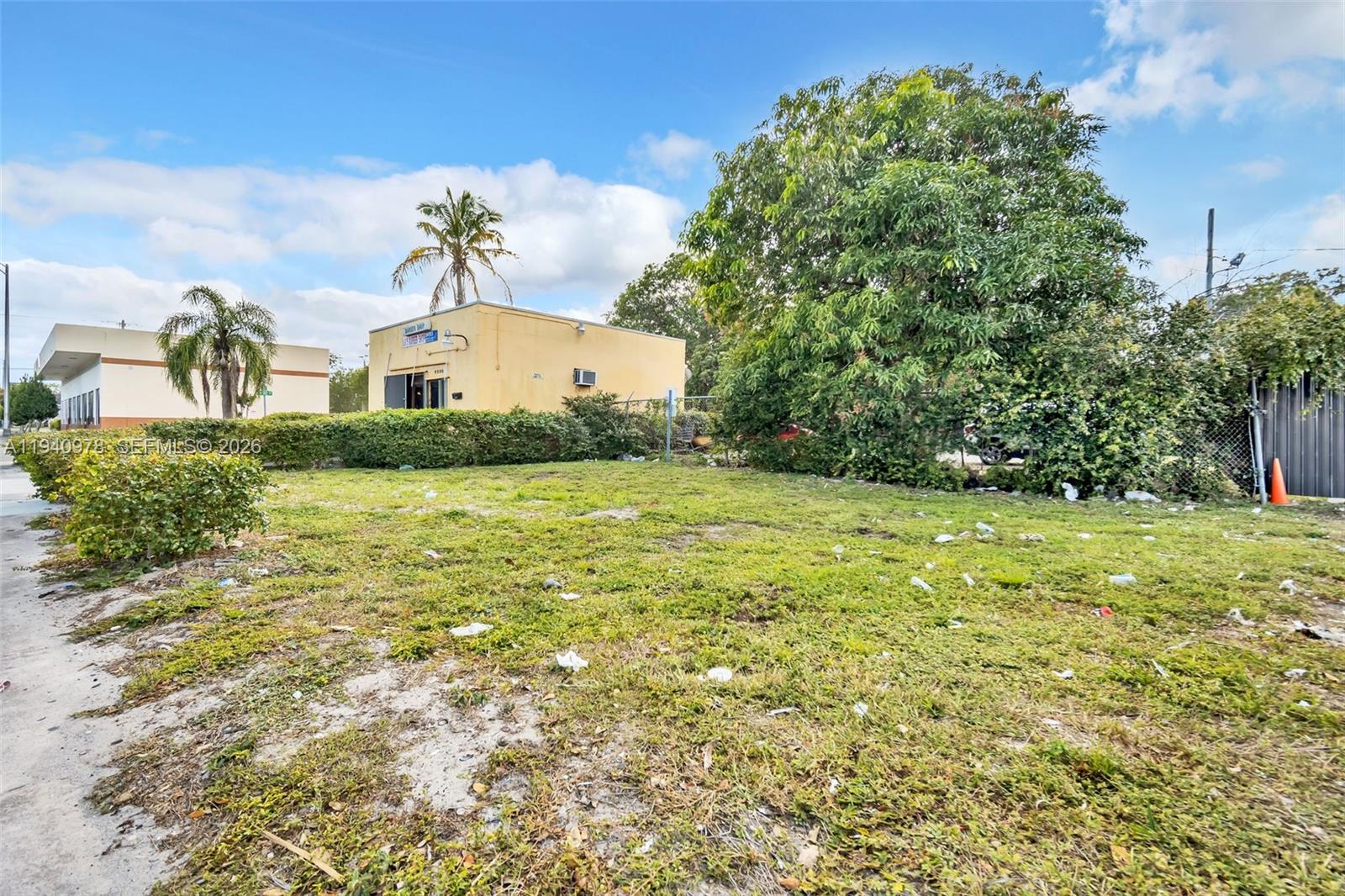 A vacant, grassy lot sits in front of a yellow commercial building under a blue sky — 9200 NW 22nd Ave, Miami FL 33147, MIAMI, FL