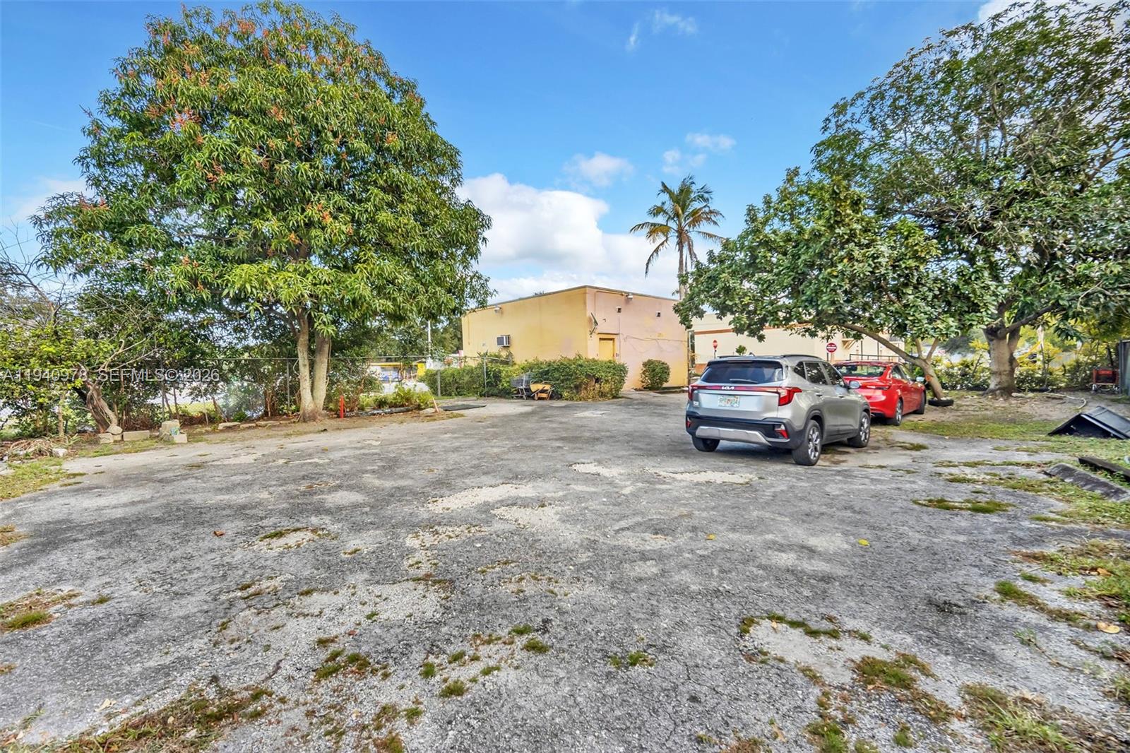 An outdoor parking area features a weathered asphalt surface, two parked cars, and mature trees — 9200 NW 22nd Ave, Miami FL 33147, MIAMI, FL