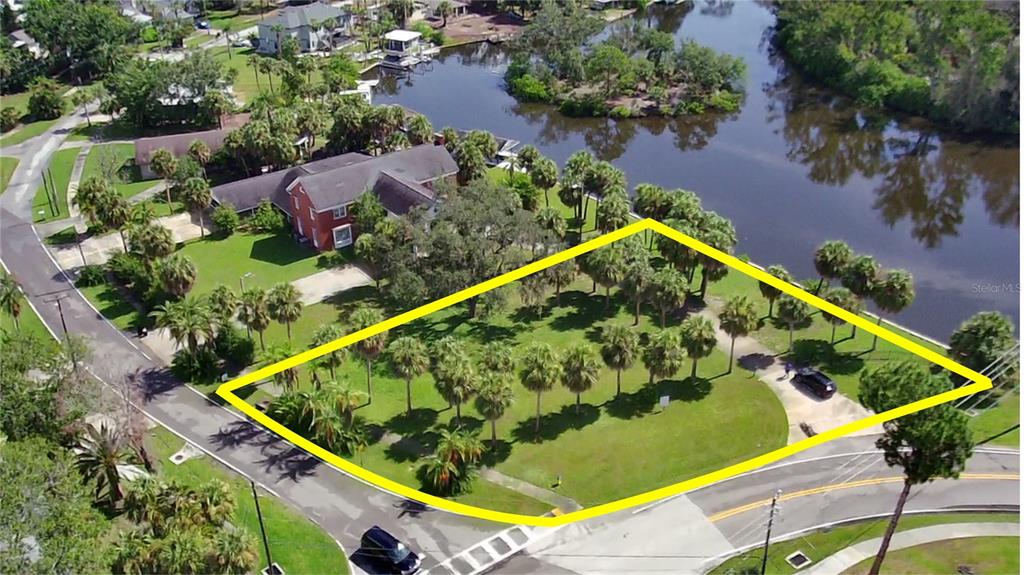 An aerial view shows a vacant, palm-tree-filled lot outlined in yellow next to a river — PARK AVENUE, TARPON SPRINGS, FL