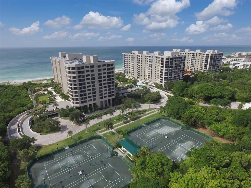 An aerial view shows two high-rise apartment buildings near the ocean with tennis courts below — 1211 GULF OF MEXICO DRIVE UNIT 1005, LONGBOAT KEY, FL