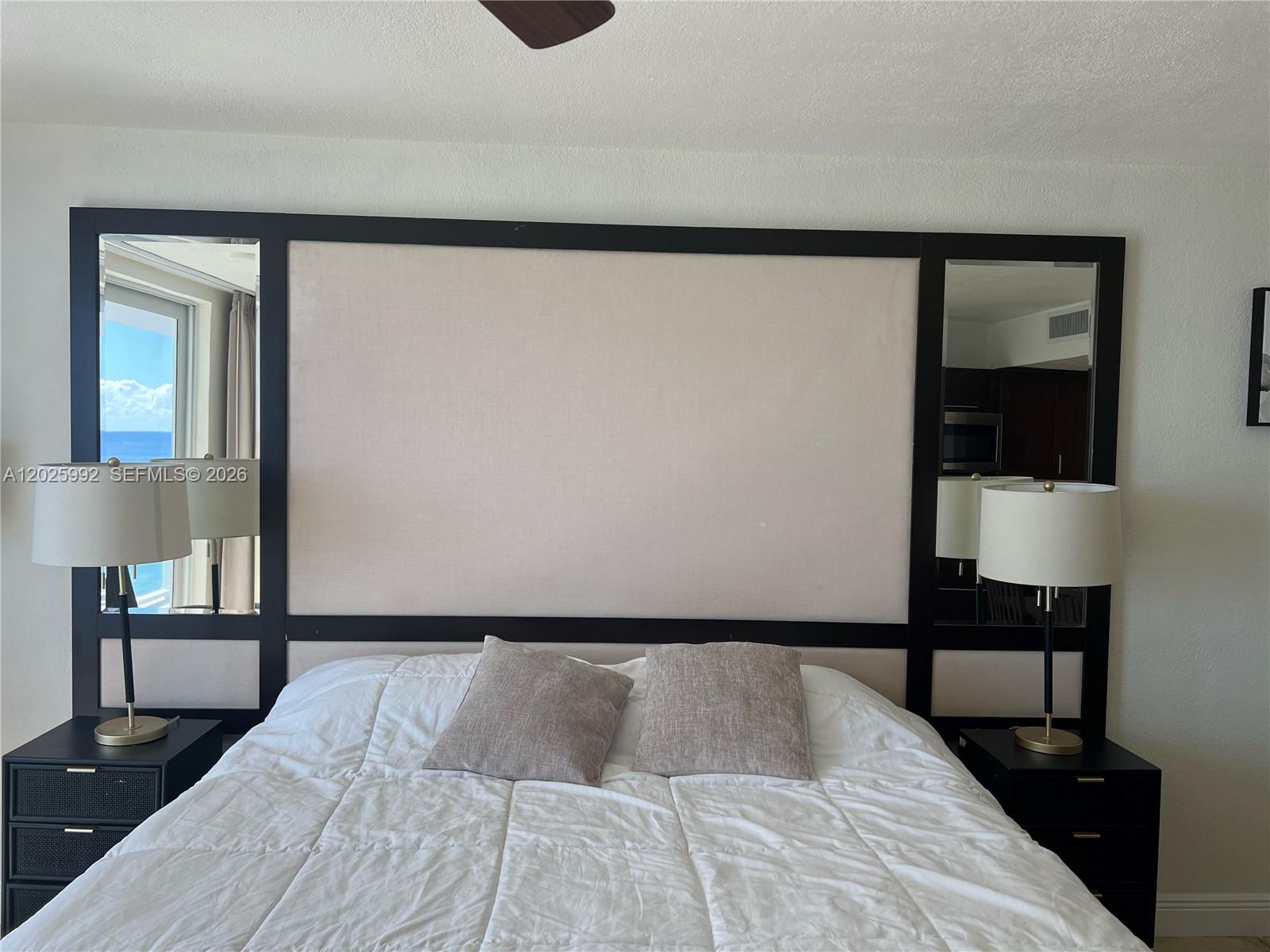 A bedroom features a large headboard with integrated mirrors and matching black nightstands with lamps — SUNNY ISLES BEACH, FL