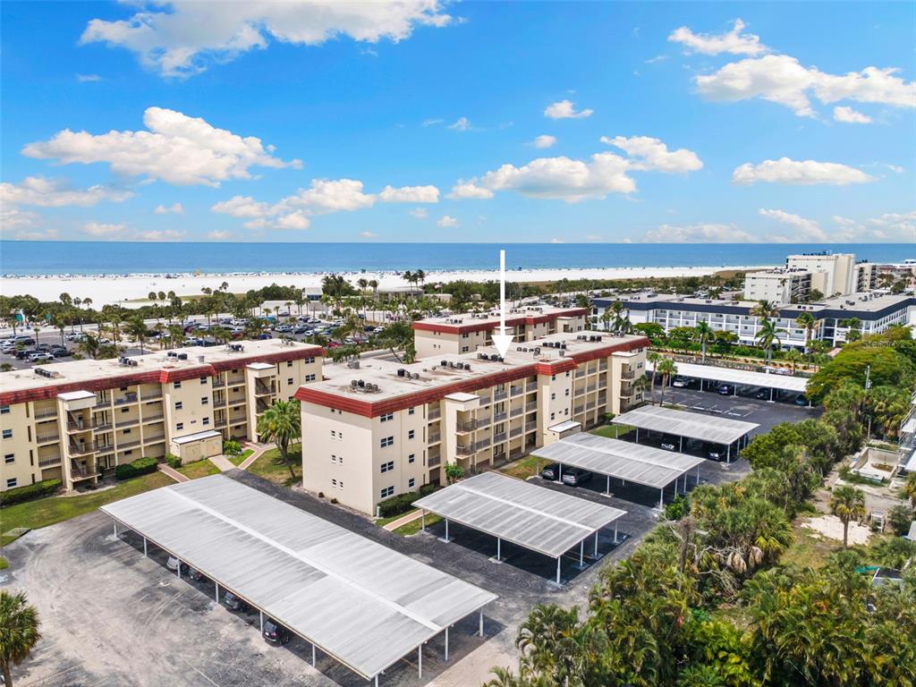 An aerial view shows multi-story apartment buildings with covered parking lots near a sandy beach — 915 BEACH ROAD UNIT 318, SARASOTA, FL