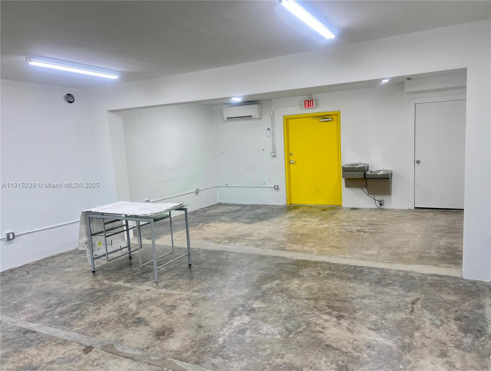 An empty room features concrete floors, white walls, a yellow exit door, and a metal table — 3070 NW 46th St, Miami FL 33142, MIAMI, FL