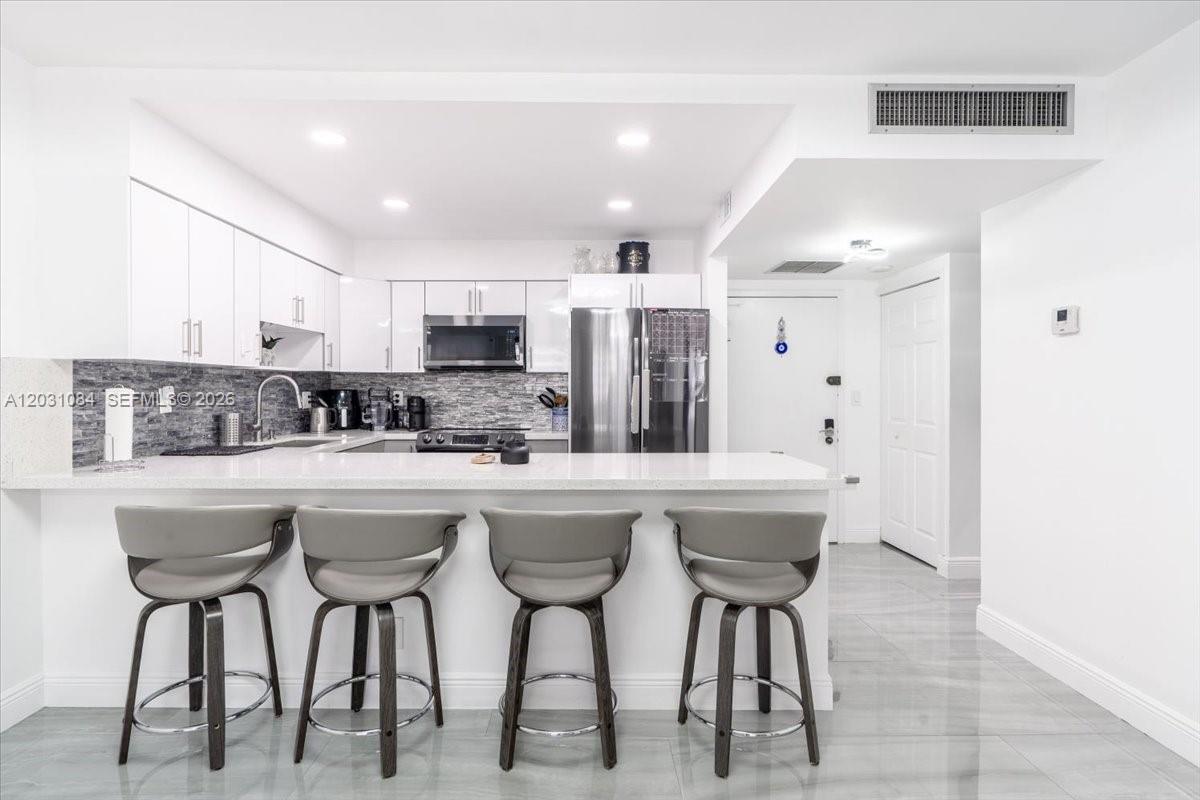 A modern kitchen features white cabinetry, stainless steel appliances, and a breakfast bar with stools — 600 Three Islands Blvd # 1413, Hallandale Beach FL 33009, HALLANDALE BEACH, FL