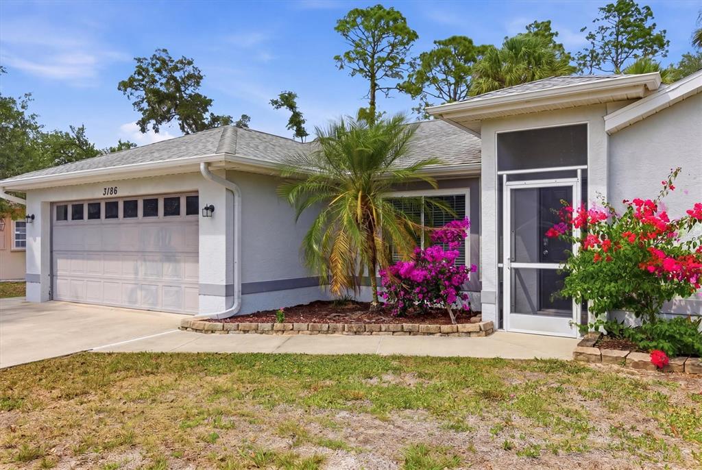 A single-story house exterior features a two-car garage, a screened entryway, and flowering landscaping — 3186 MARITA AVENUE, NORTH PORT, FL