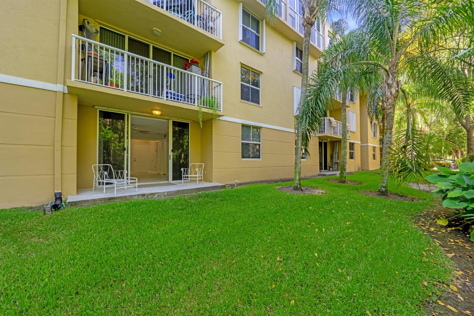 A ground-floor apartment patio with white metal chairs sits beside a grassy lawn and palms — 619 E Sheridan Street # 106, Dania Beach FL 33004, DANIA BEACH, FL