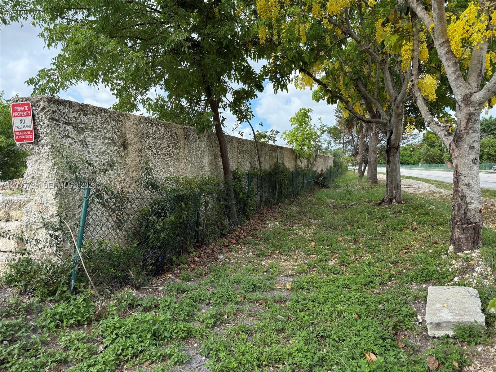 A long, weathered concrete wall runs alongside a chain-link fence and trees near a road — SW 157th, Homestead FL 33033, HOMESTEAD, FL