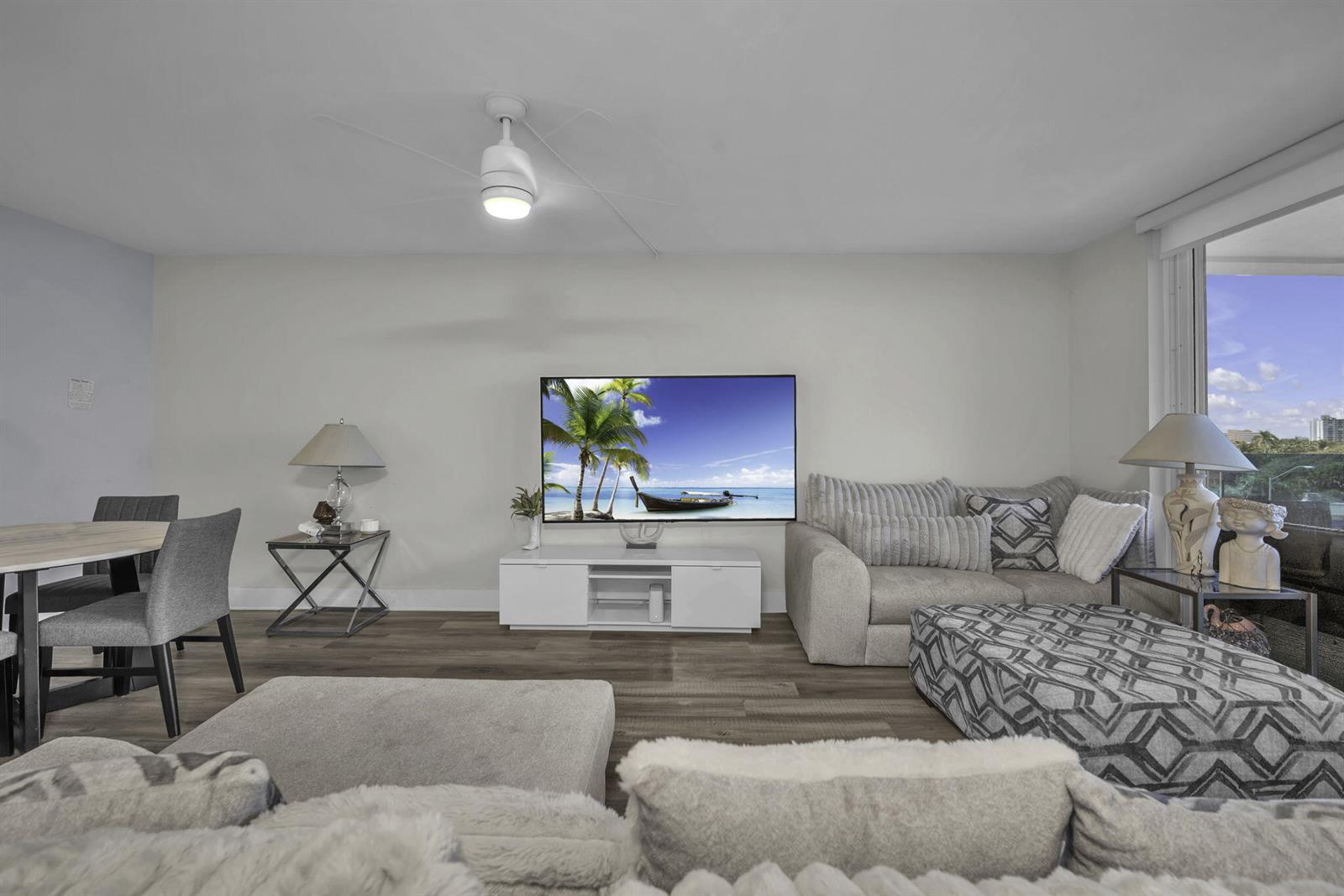 A living room features a wall-mounted television, gray sofa, patterned ottoman, and wood-look flooring — 2639 N Riverside Drive # 505, Pompano Beach FL 33062, POMPANO BEACH, FL