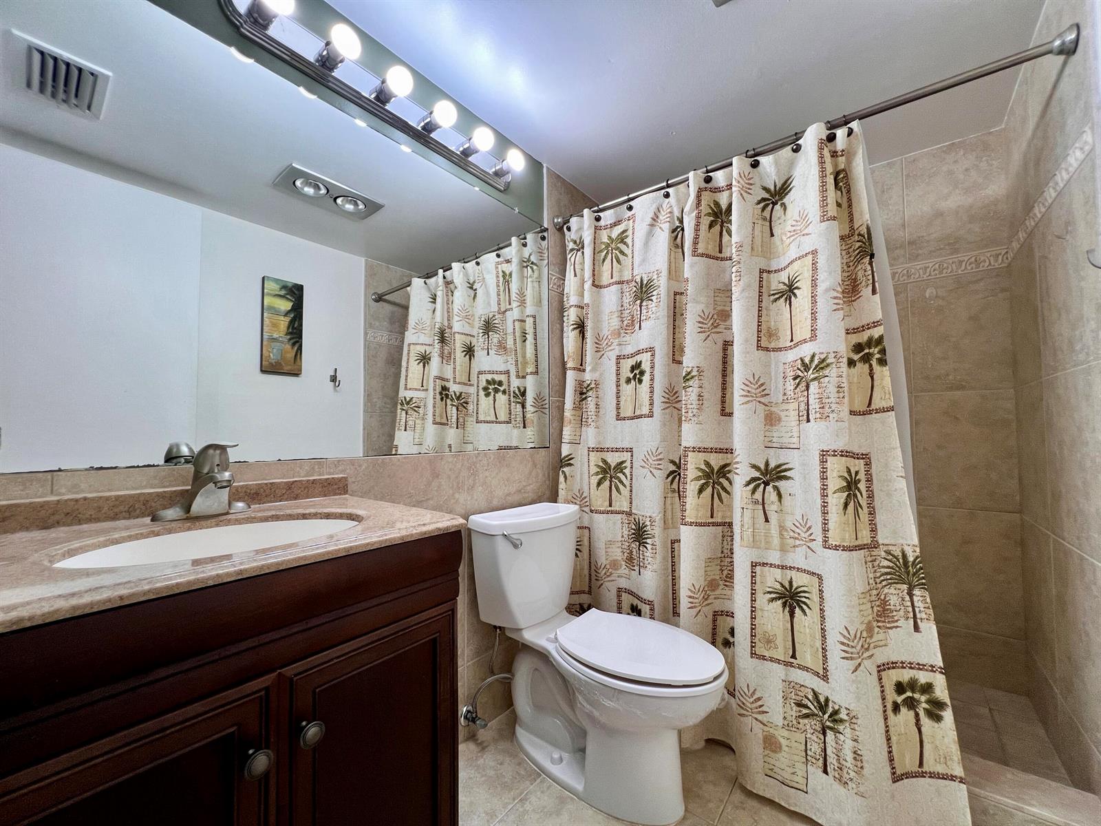 A bathroom features a dark wood vanity, beige tile walls, and a palm-print shower curtain — 8 Northampton A, West Palm Beach FL 33417, WEST PALM BEACH, FL