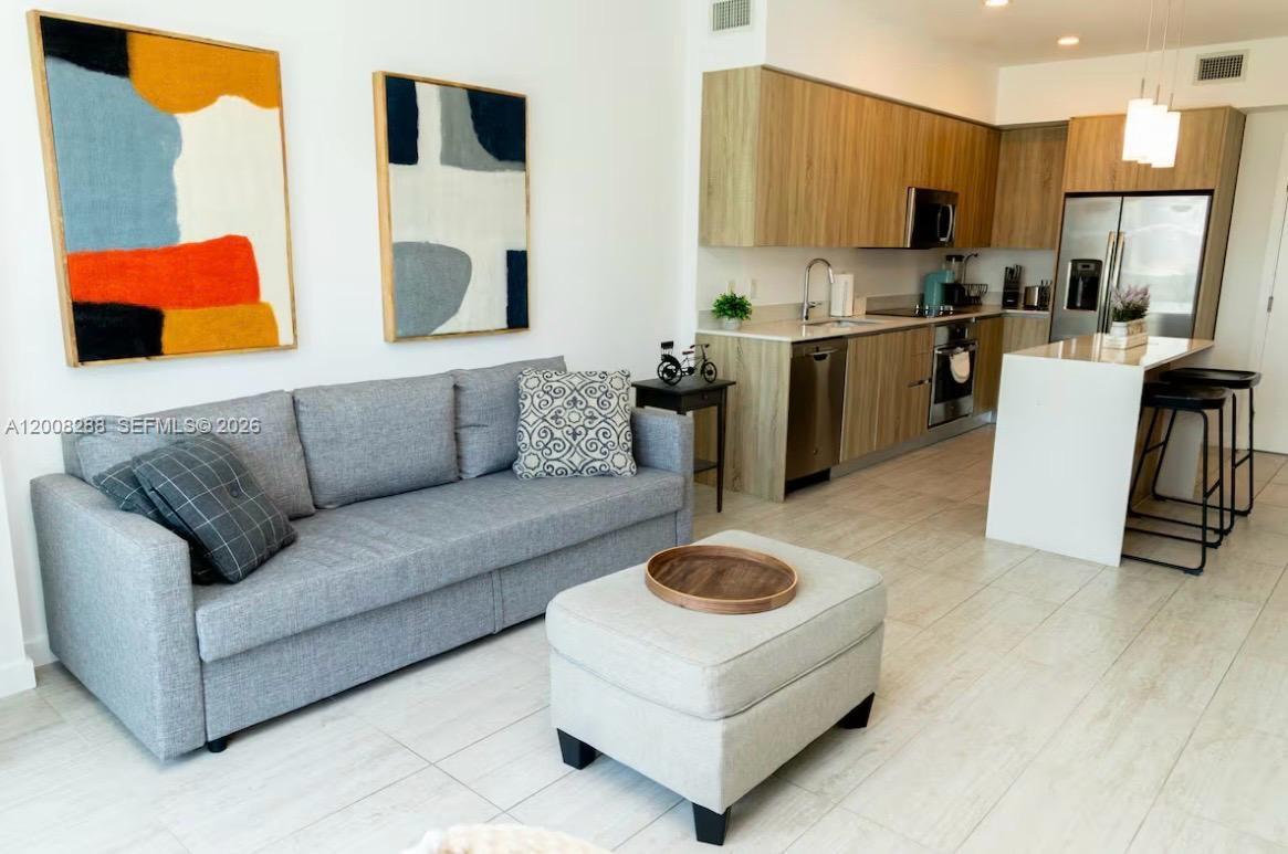 A modern living room features a grey sofa, an ottoman, and an open-concept kitchen — 3900 Biscayne Blvd # 906, Miami FL 33137, MIAMI, FL
