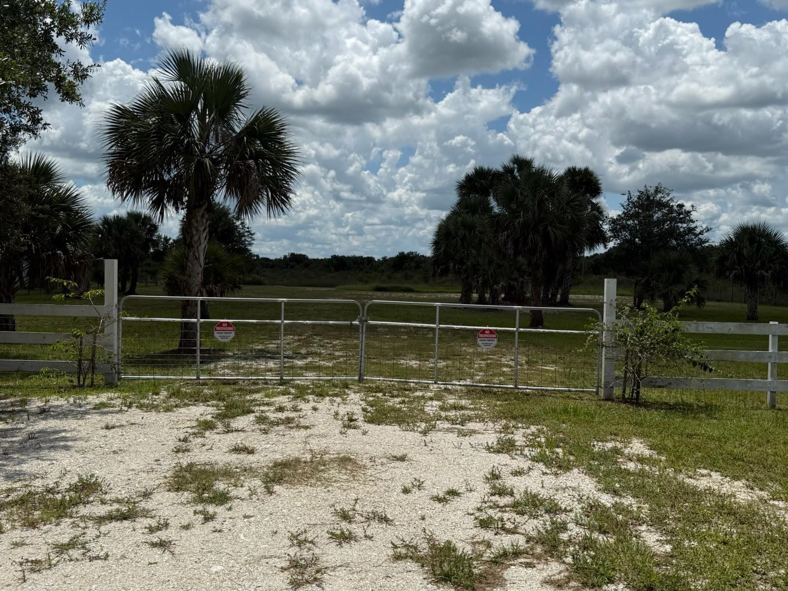 A white fence and metal gate enclose a gravel driveway leading to a grassy field — 19374 NW 302, Okeechobee FL 34972, OKEECHOBEE, FL