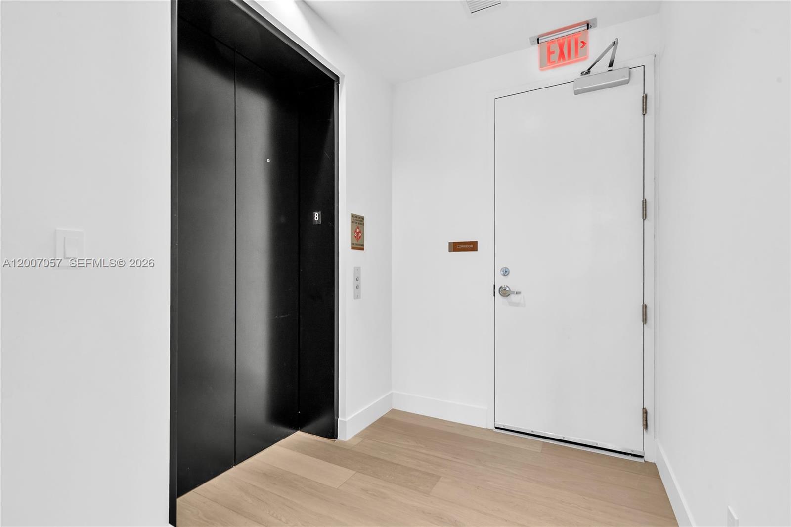 A modern elevator lobby features black elevator doors, a white exit door, and light wood flooring — 175 SE 25 Road # 803, Miami FL 33129, MIAMI, FL