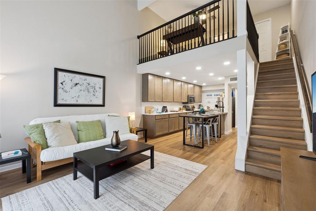 A living room with a futon, coffee table, and kitchen area beneath a lofted balcony — 6168 PEREGRINE AVENUE UNIT 12B, ORLANDO, FL