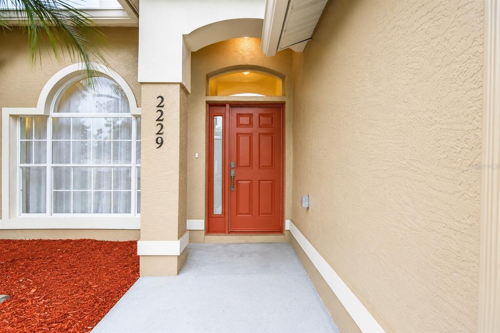 A red front door with a side window sits under a covered entryway — 2229 FOGGY RIDGE PARKWAY, LAND O LAKES, FL