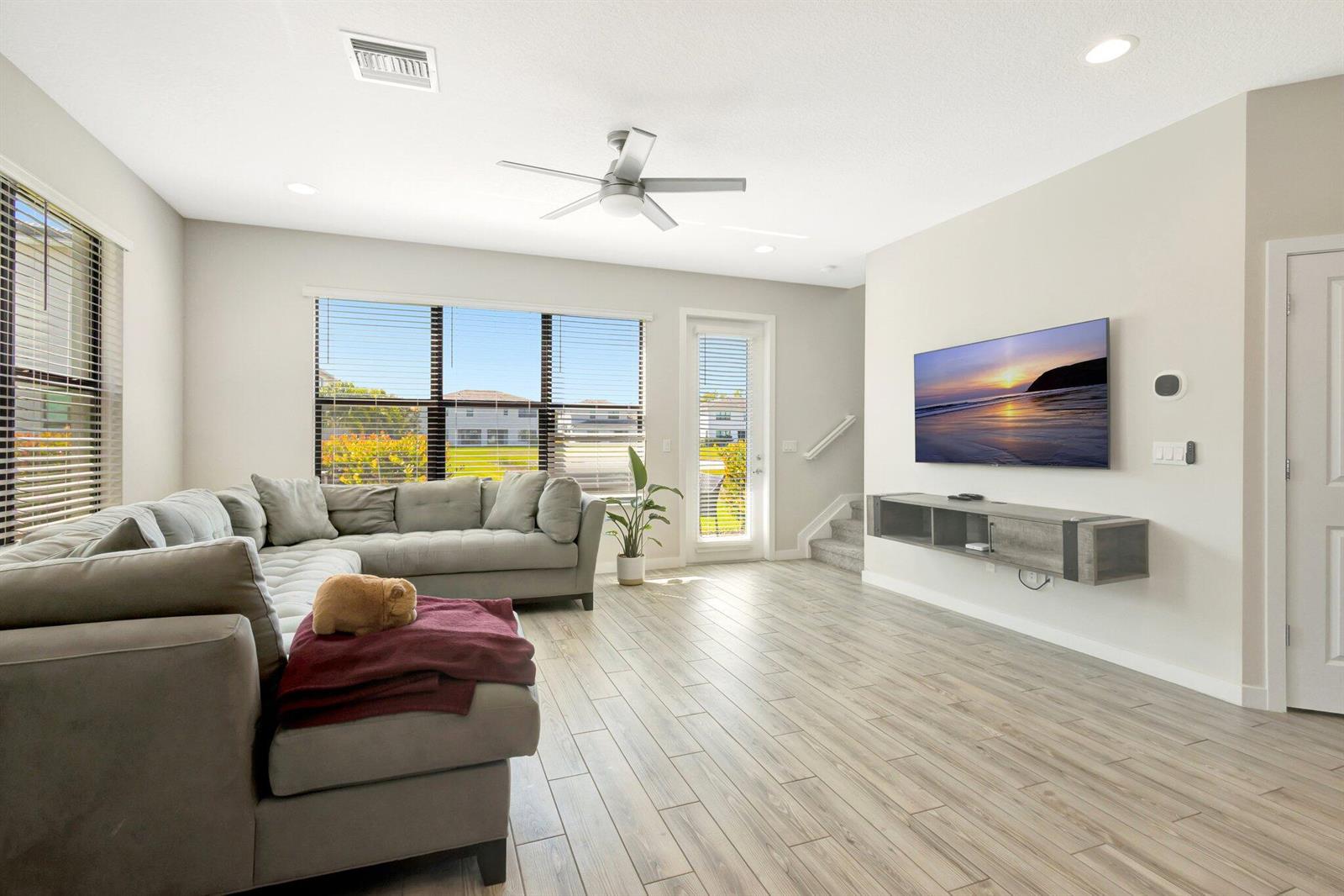 A living room features a grey sectional sofa, a wall-mounted television, and light wood flooring — 8654 Antarus Drive, Lake Worth FL 33467, LAKE WORTH, FL