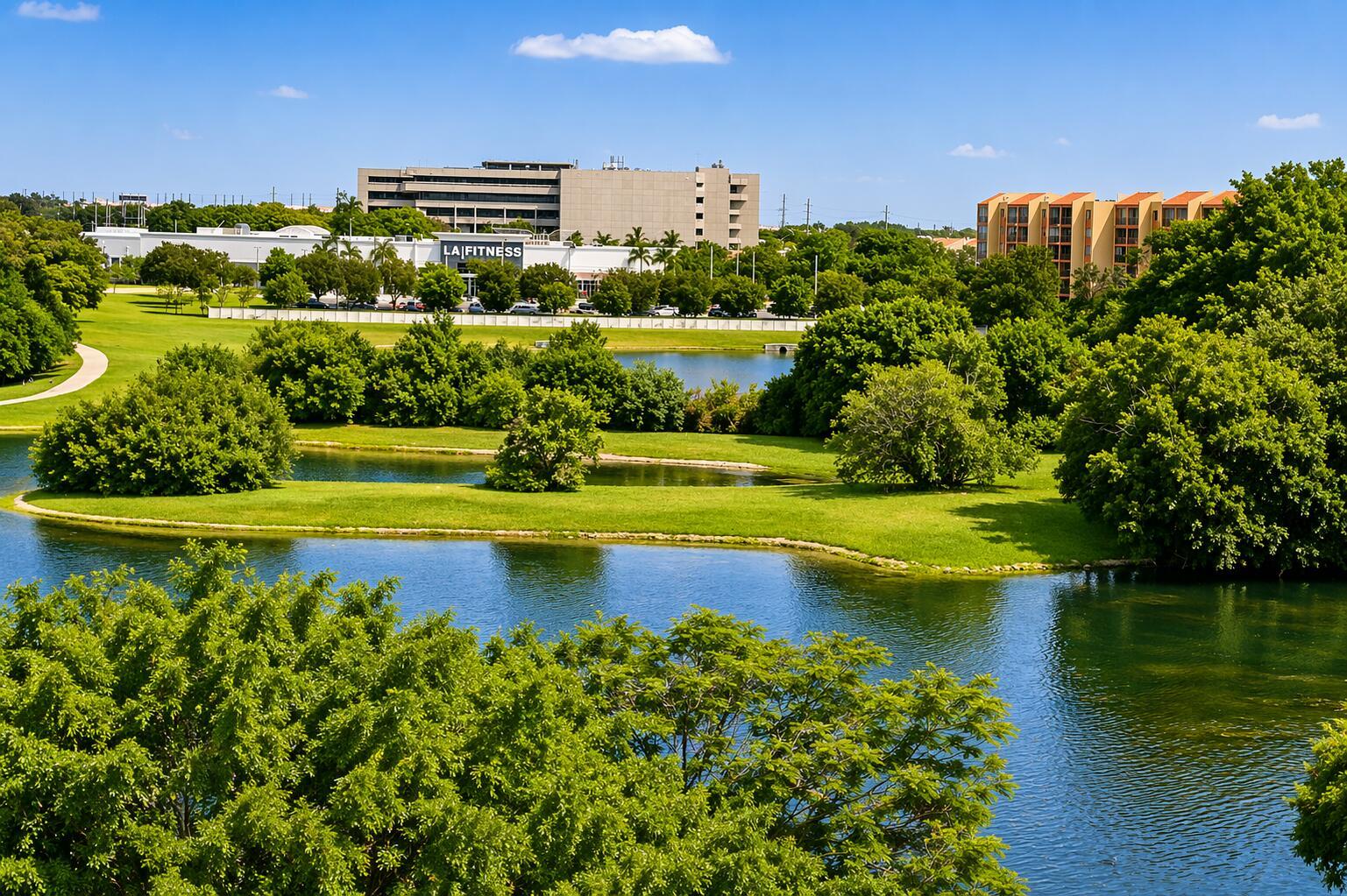 An outdoor view of a pond surrounded by green trees and nearby commercial buildings — 9682 Fontainebleau Boulevard # 603, Miami FL 33172, MIAMI, FL