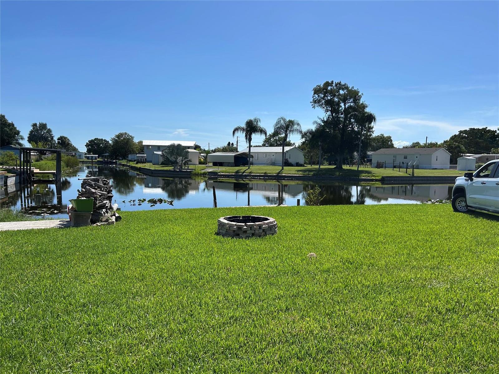 A grassy backyard features a stone fire pit overlooking a canal with houses beyond — 1379 Chobee Street, Okeechobee FL 34974, OKEECHOBEE, FL