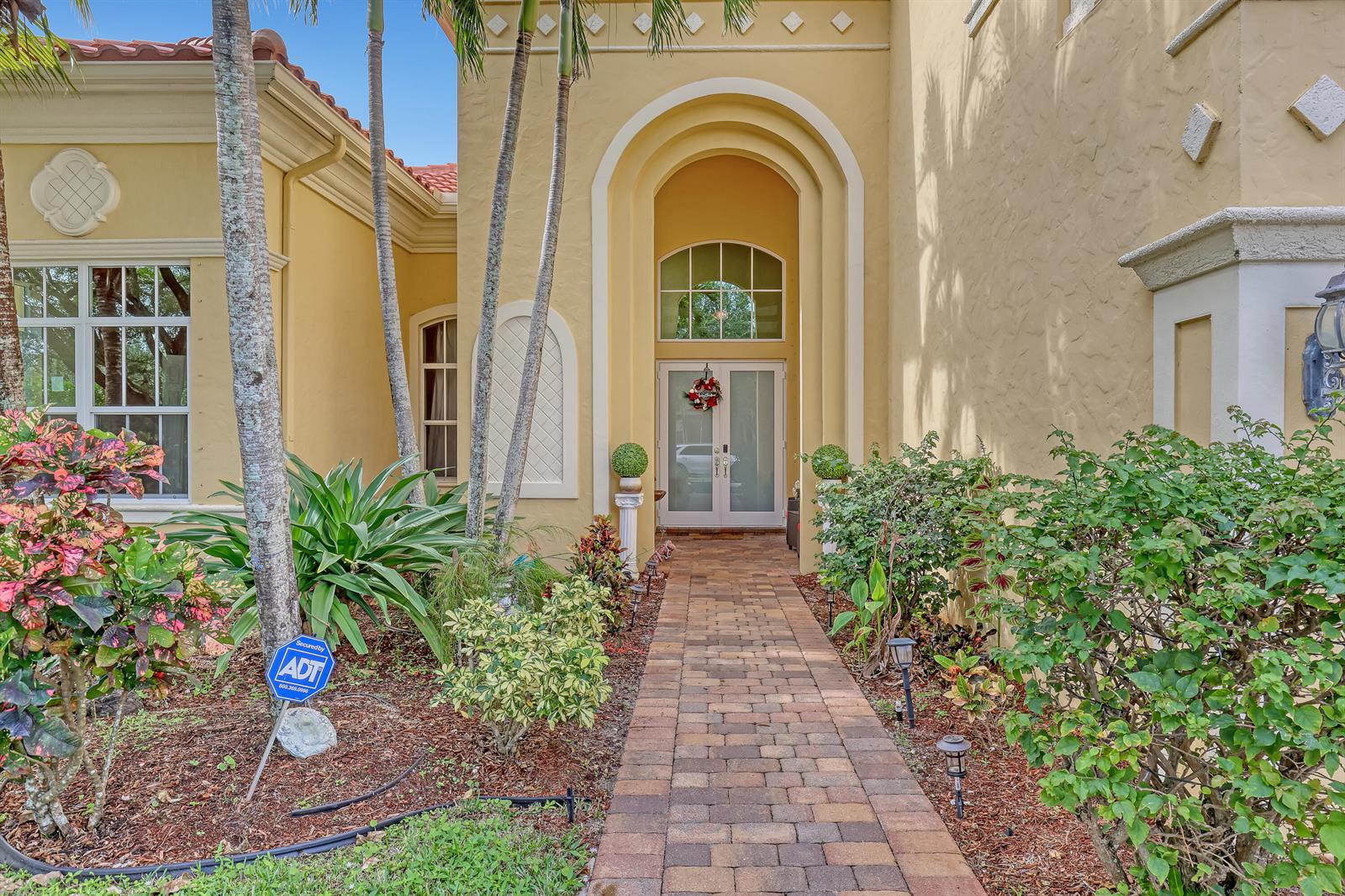 A paved walkway leads to the arched entryway of a yellow stucco house with double doors — 3389 Lago De Talavera, Lake Worth FL 33467, LAKE WORTH, FL