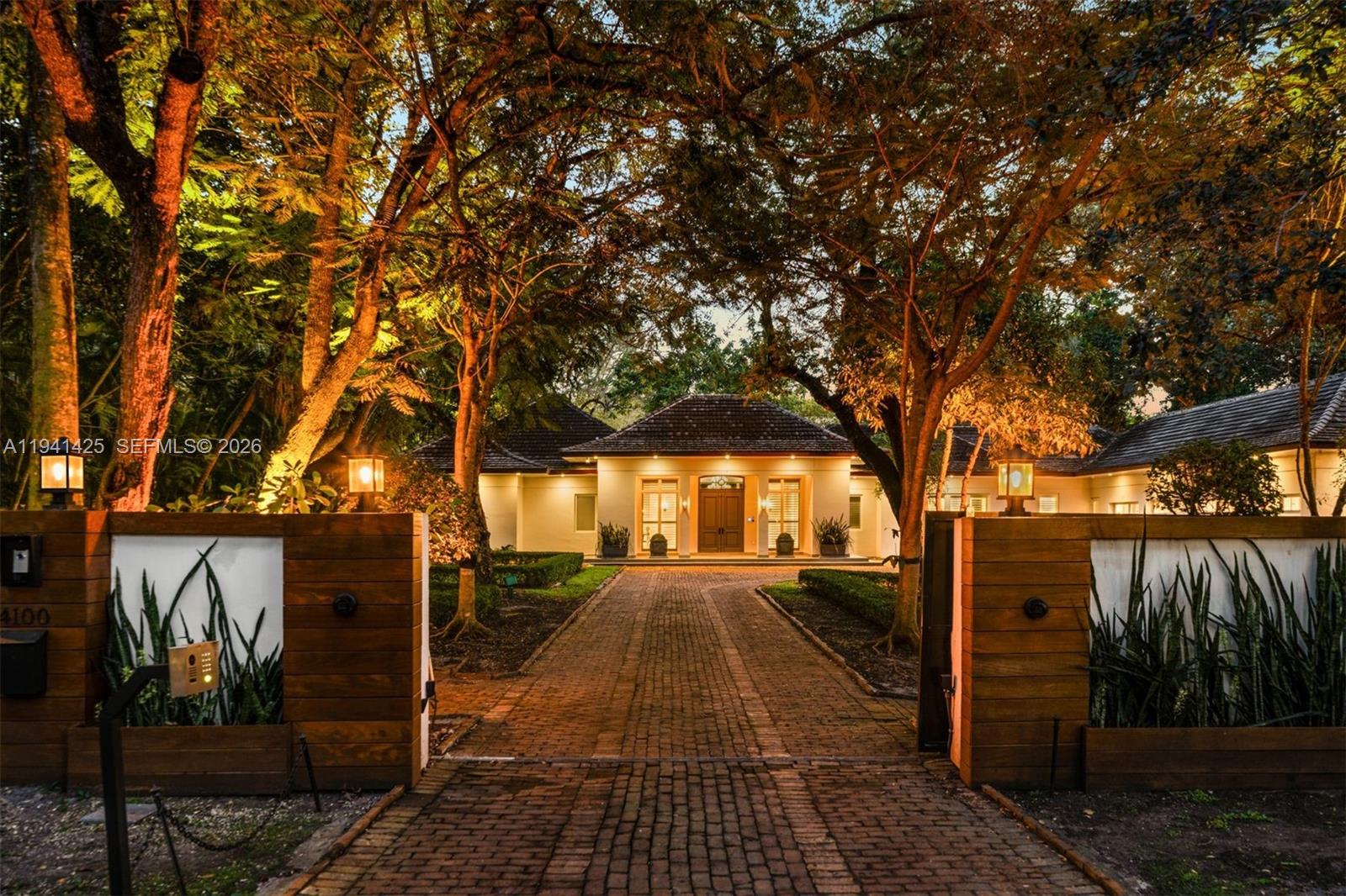 A brick driveway leads to a single-story house framed by large trees and gated walls — 4100 Kiaora St, Miami FL 33133, MIAMI, FL
