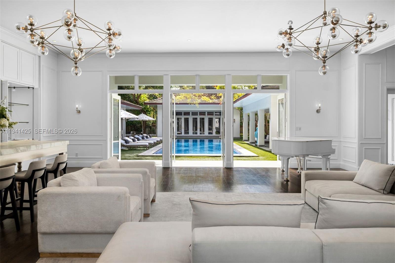 A living room with two chandeliers, white furniture, a piano, and a view of a pool — 4100 Kiaora St, Miami FL 33133, MIAMI, FL