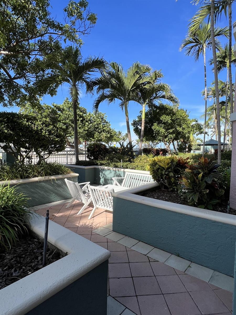 An outdoor patio area features white chairs, raised garden beds, and palm trees under blue skies — 2051 SE 3rd Street, Deerfield Beach FL 33441, DEERFIELD BEACH, FL