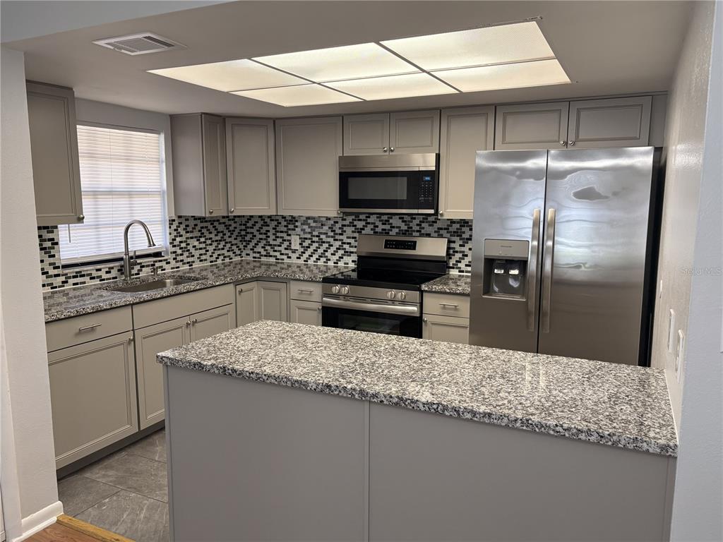 A kitchen features gray cabinets, granite countertops, a mosaic tile backsplash, and stainless steel appliances — 4015 ASHFORD GREEN PLACE UNIT D202, TAMPA, FL