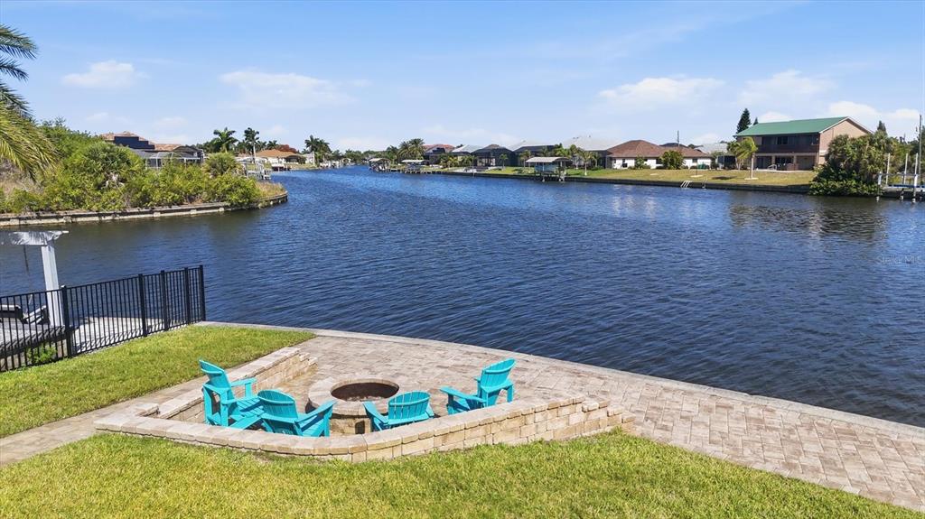 A stone patio with a fire pit and blue chairs overlooks a residential canal — 9266 MIGUE CIRCLE, PORT CHARLOTTE, FL