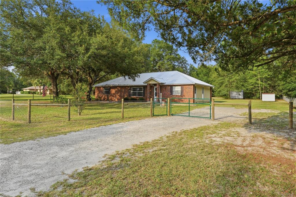 A single-story brick house with a metal roof sits behind a fenced gravel driveway — 5480 NE 53RD TERRACE, HIGH SPRINGS, FL