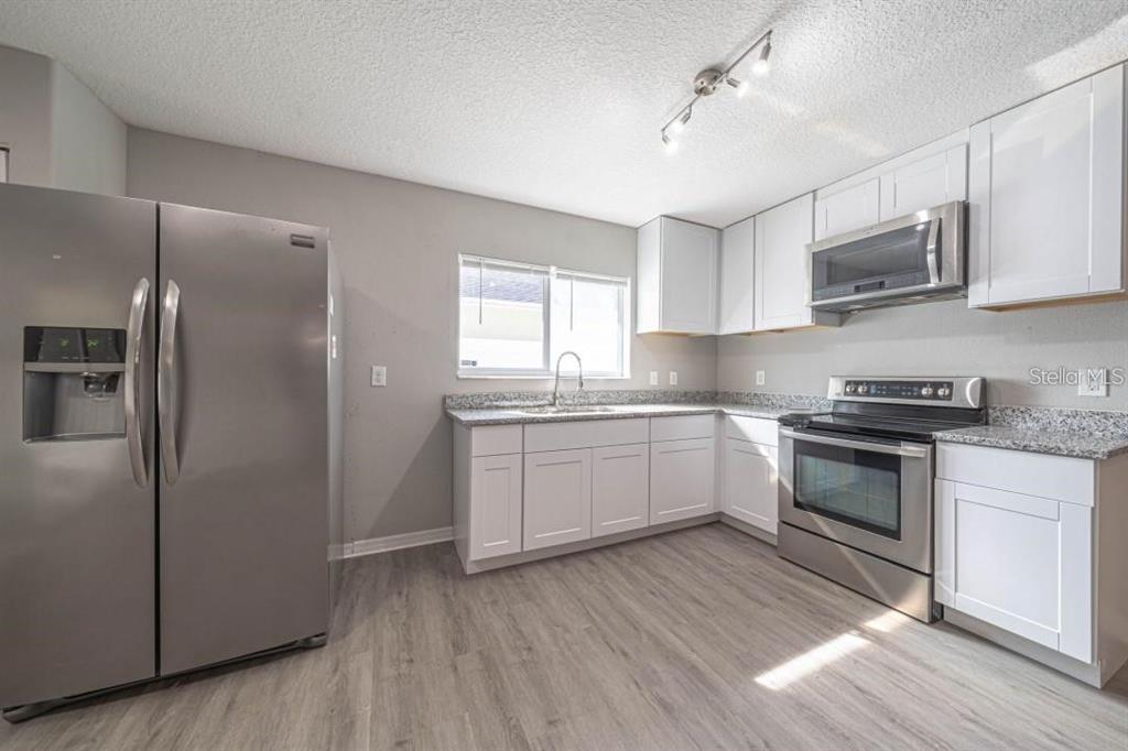 A kitchen features white cabinets, stainless steel appliances, granite countertops, and light wood-look flooring — 2025 PEACEFUL PALM STREET, RUSKIN, FL