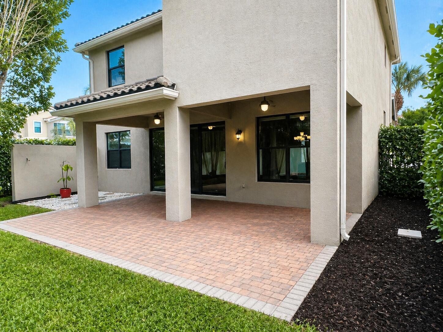 A covered patio with brick pavers sits beneath the second story of a beige house — 8079 Snowflake Obsidian Trail, Delray Beach FL 33446, DELRAY BEACH, FL