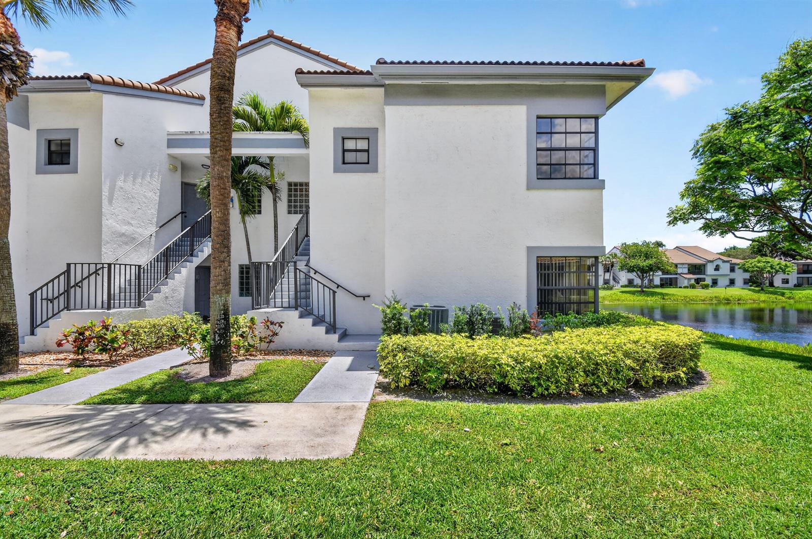 A two-story white apartment building with exterior stairs sits beside a pond and lawn — 5394 Firenze Drive, Boynton Beach FL 33437, BOYNTON BEACH, FL