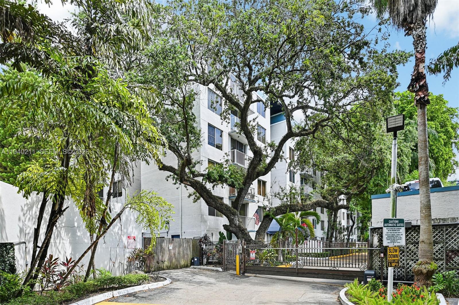 A gated entrance leads to a multi-story white apartment building surrounded by large trees — 1740 NW N River Dr # 614, Miami FL 33125, MIAMI, FL