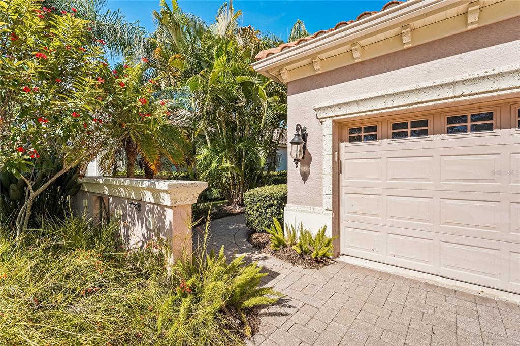 A beige garage door with windows sits beside a stone walkway and lush tropical landscaping — 7216 PRESIDIO GLEN, LAKEWOOD RANCH, FL