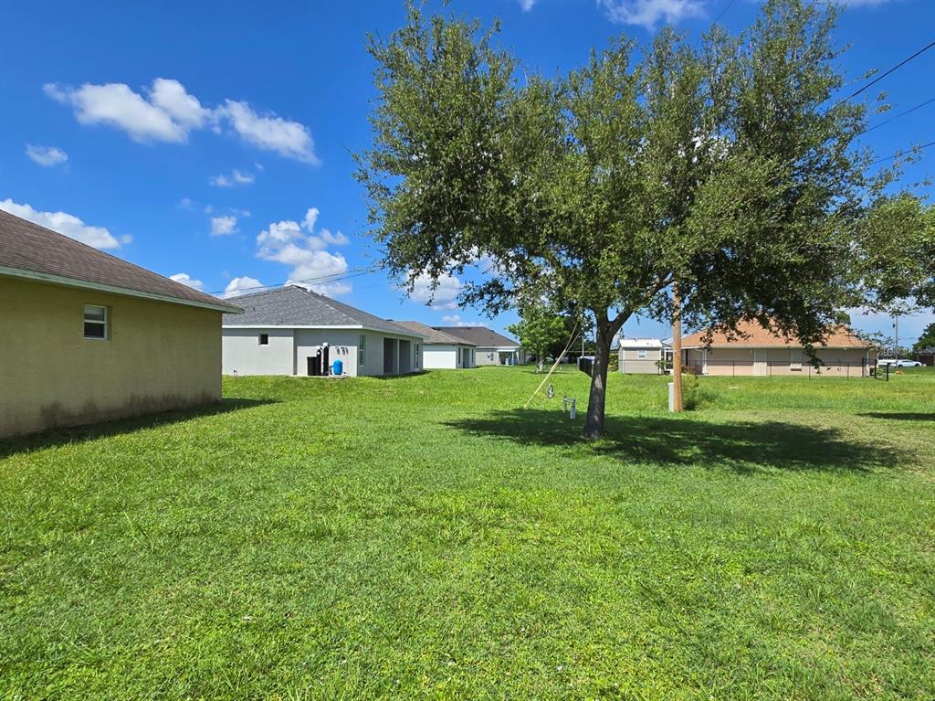 A grassy backyard with a large tree and several neighboring houses under a blue sky — 519 NW 26TH STREET, CAPE CORAL, FL