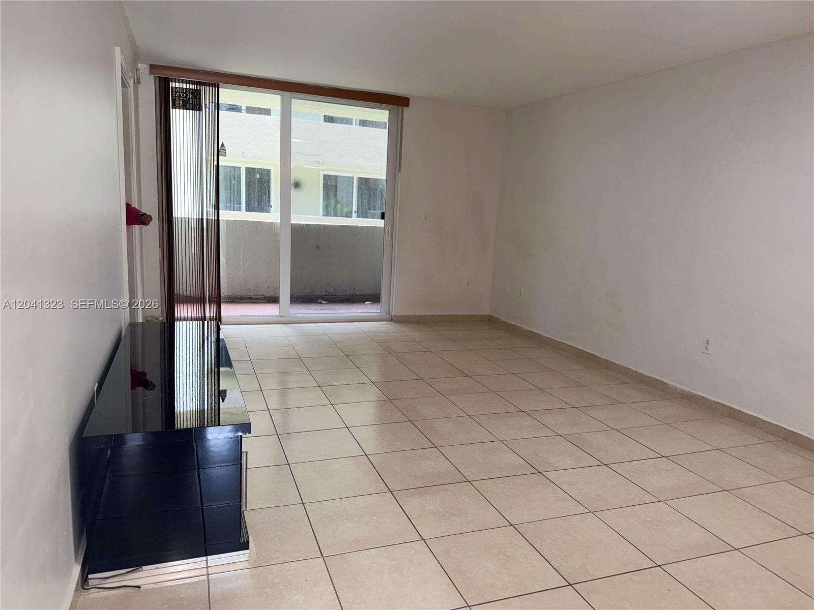 An empty living room features beige tile flooring, white walls, and a sliding glass door — 10090 NW 80th Ct # 1241, Hialeah Gardens FL 33016, HIALEAH GARDENS, FL
