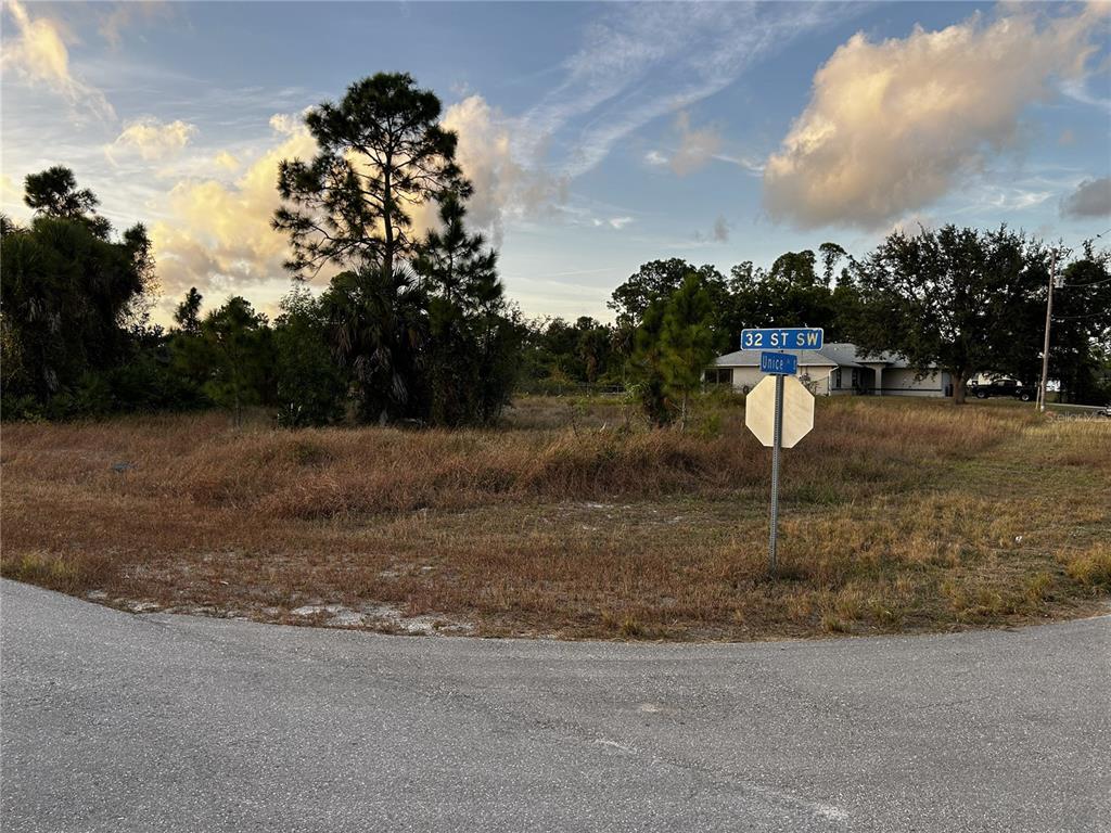 A vacant, grassy corner lot features a street sign and scattered trees under a cloudy sky — 3506 32ND STREET SW, LEHIGH ACRES, FL