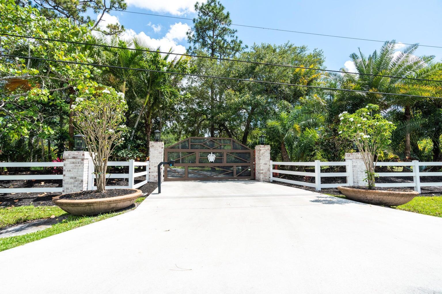 A concrete driveway leads to a gated entrance flanked by white fences and potted trees — 10780 Anderson Lane, Wellington FL 33449, WELLINGTON, FL