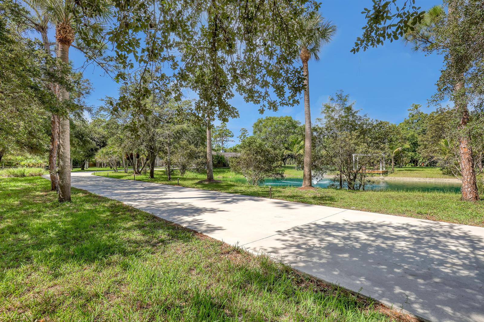 A concrete driveway curves through a grassy yard toward a pond with a fountain — 10780 Anderson Lane, Wellington FL 33449, WELLINGTON, FL