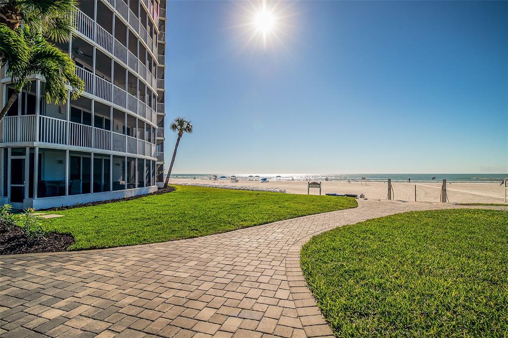 A paved walkway leads past a multi-story apartment building toward a sunny sandy beach — 6300 MIDNIGHT PASS ROAD UNIT 21, SARASOTA, FL