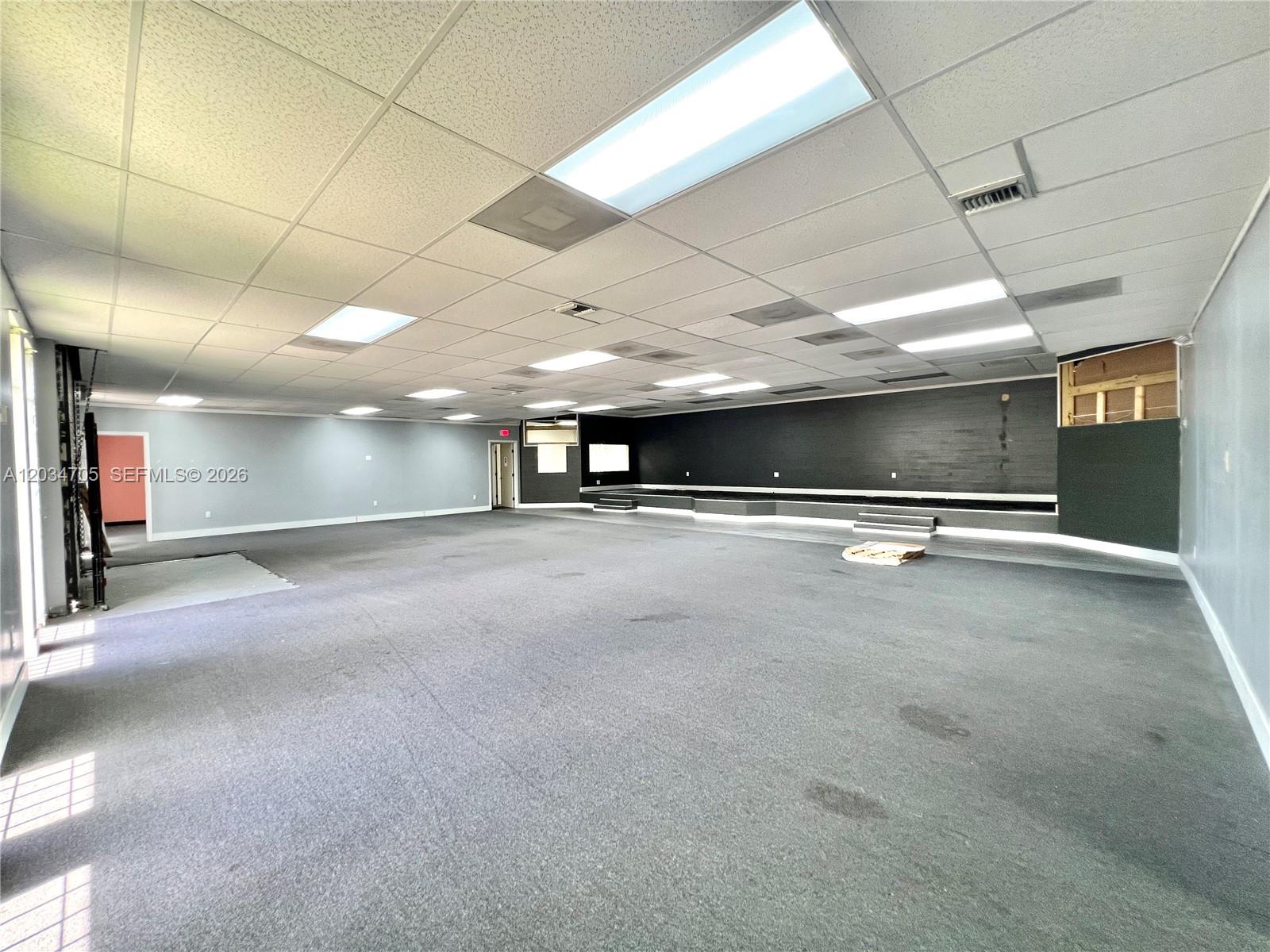 A large, empty commercial space features gray carpeting, a drop ceiling, and a raised stage area — 3425 NW 73rd Ave, Miami FL 33122, MIAMI, FL