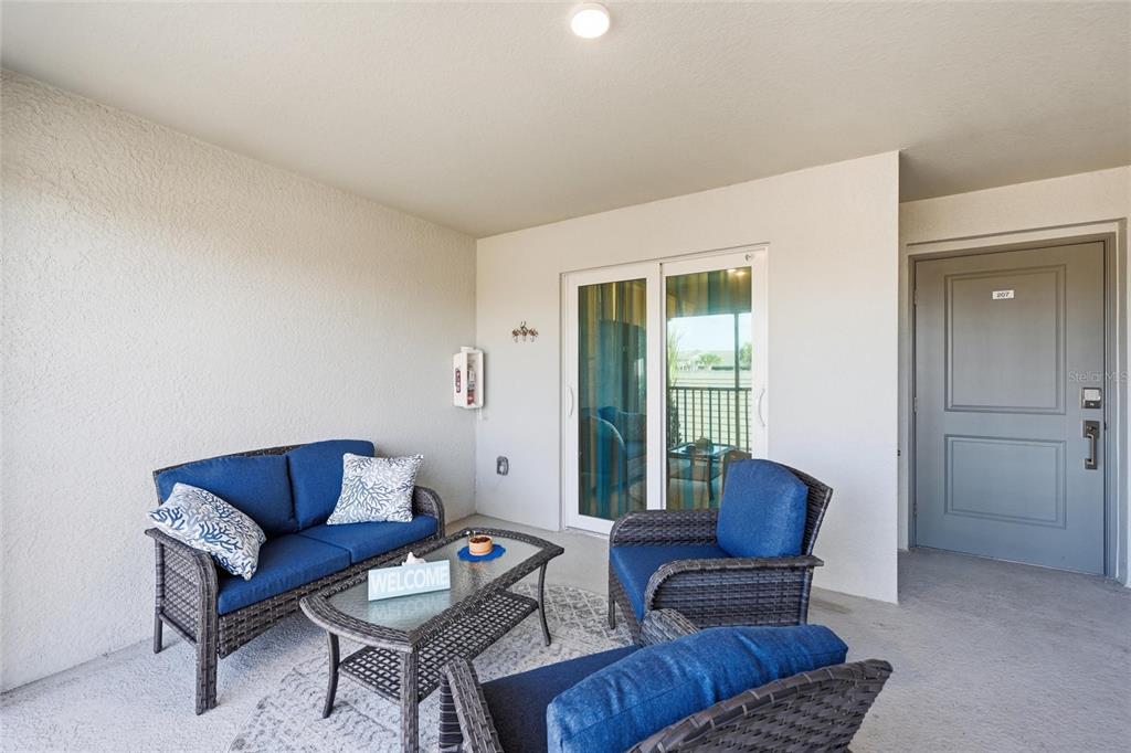 A covered balcony features blue wicker furniture, a glass-topped coffee table, and a gray door — 17505 OPAL SAND COURT UNIT 207, VENICE, FL