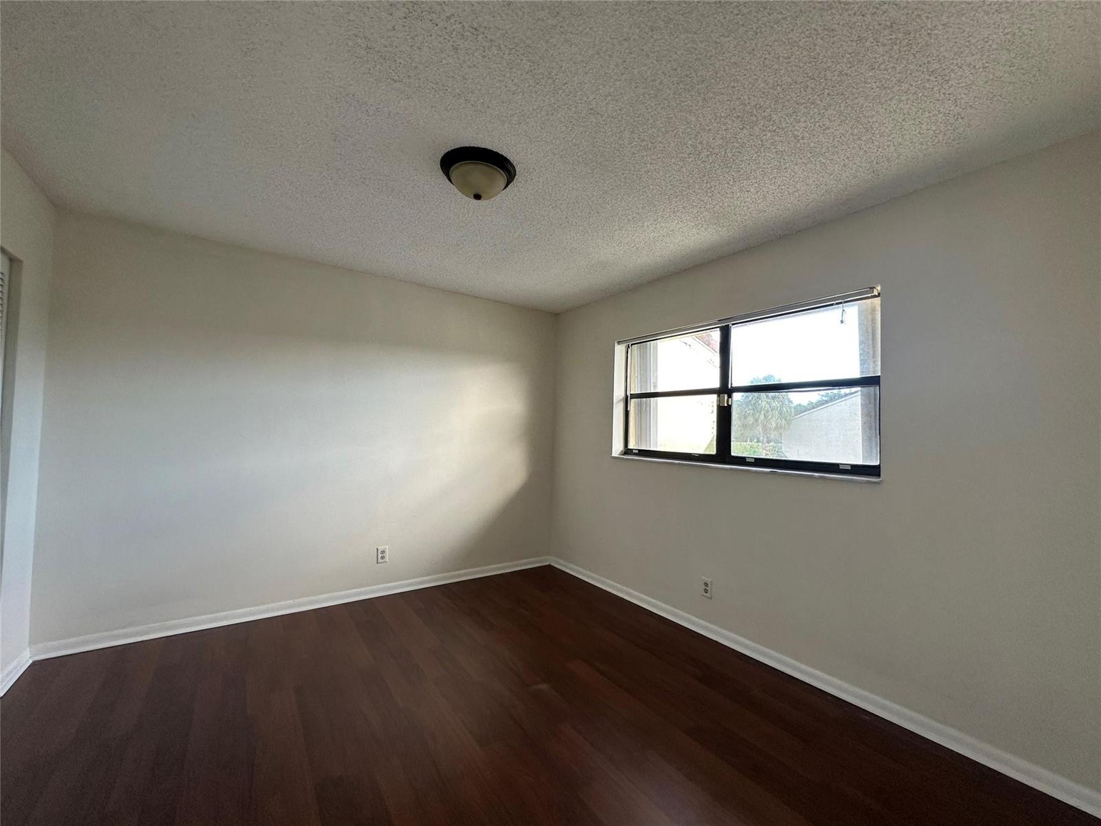 An empty bedroom features white walls, dark wood-look flooring, a popcorn ceiling, and a window — 22415 SW 61st Way # 202, Boca Raton FL 33428, BOCA RATON, FL