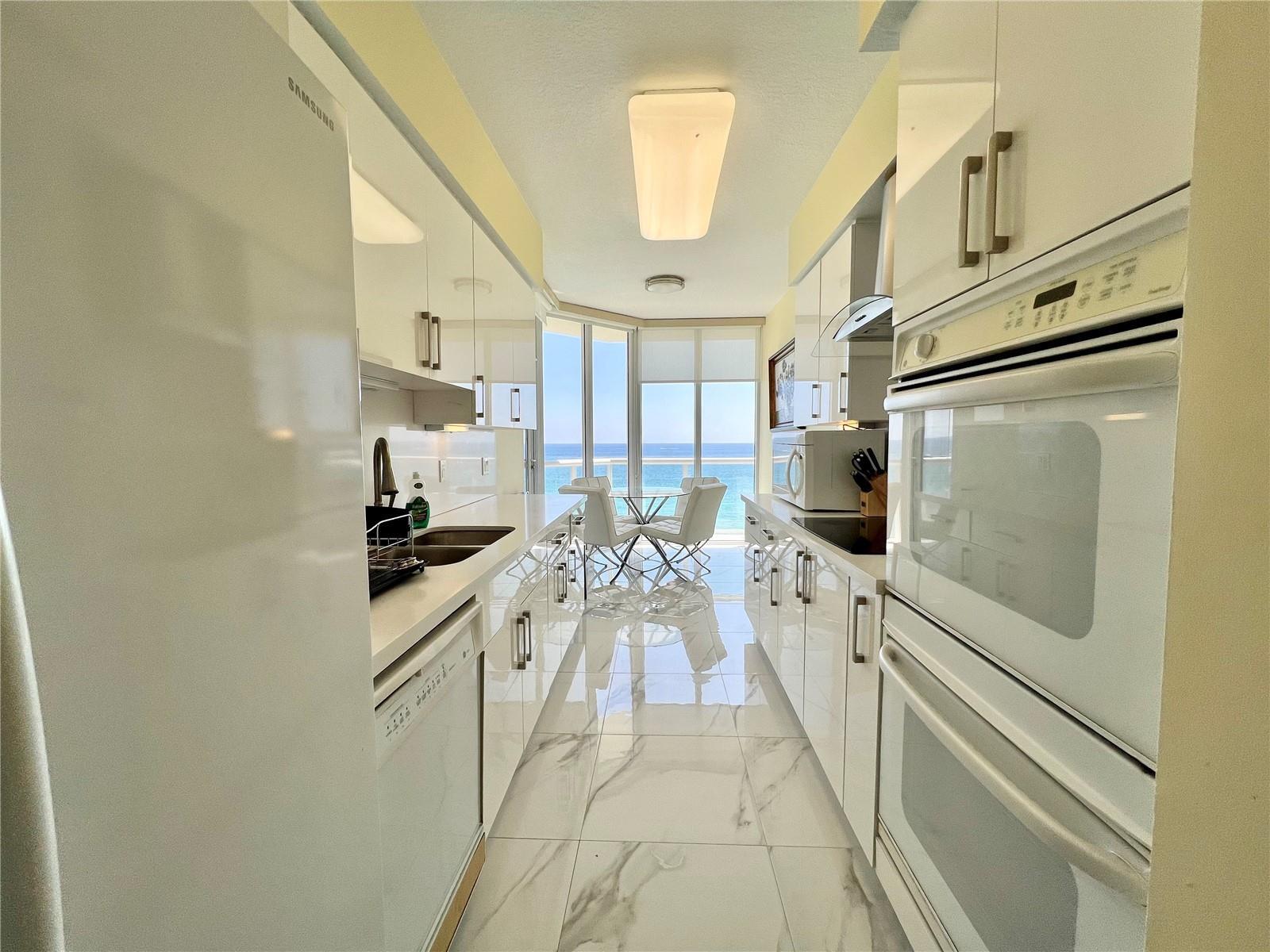 A galley kitchen features white glossy cabinets, marble-look tile floors, and a view of the ocean — 6001 N Ocean Drive # 706, Hollywood FL 33019, HOLLYWOOD, FL