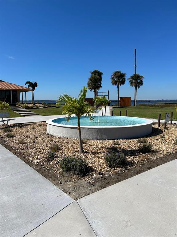 A circular fountain sits in a gravel landscape with palm trees near a waterfront building — 709 BELMONTE DRIVE, INDIAN LAKE ESTATES, FL