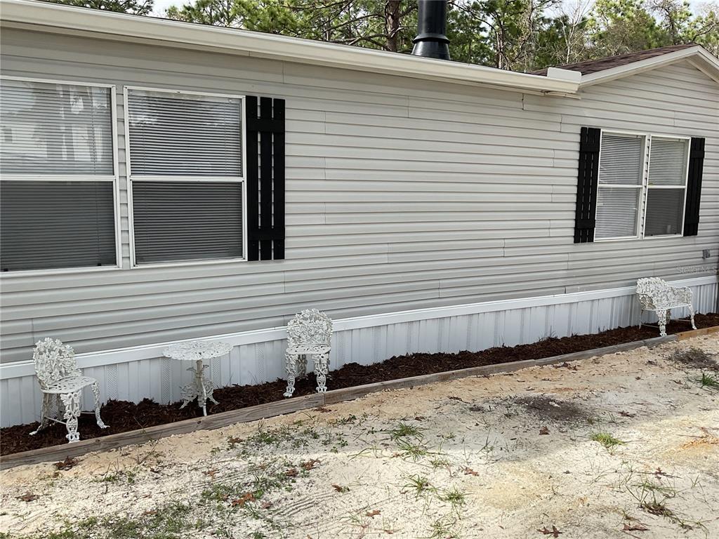 The exterior of a light gray mobile home features white skirting, black shutters, and metal furniture — 90 NE 132ND TERRACE, WILLISTON, FL